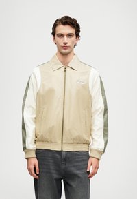 Beige Bomberjacke mit weißen und olivgrünen Ärmeln, Reißverschluss auf der Vorderseite und gerippten Strickbündchen. Mit einem Logo-Patch auf der linken Brust.