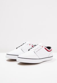 Baskets blanches en cuir texturé, semelle plate en caoutchouc, avec des accents rayés noir/rouge sur le col. Dotées de lacets traditionnels et d'un logo.