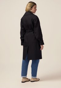 Zwarte trenchcoat met een strik taille, lange mouwen met opgerolde cuffs en een rugventilatie, gecombineerd met blauwe denim broek en beige schoenen.