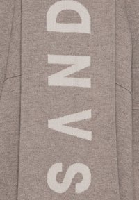 Grauer Strickpullover mit auffälligen weißen Buchstaben, die "SAND" in vertikaler Anordnung schreiben. Weiche Textur, lässiges Design und lockere Passform.