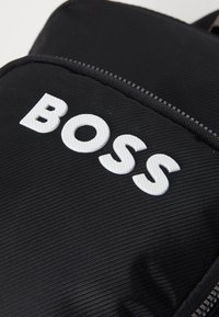 Crna najlonska torba koja ima teksturirani bijeli logo "BOSS" istaknuto prikazan. Uključuje detalj sa zatvaračem duž ruba za zatvaranje.
