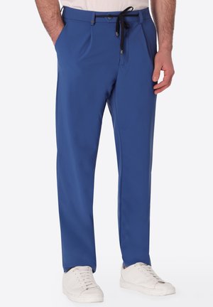 Uomo con pantaloni blu con coulisse nera e scarpe da ginnastica bianche, in piedi con le mani parzialmente nelle tasche.