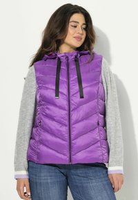 Gilet imbottito viola con cappuccio, dotato di zip nera e cordini. Motivo a chevron, abbinato a una maglietta a maniche lunghe grigia.
