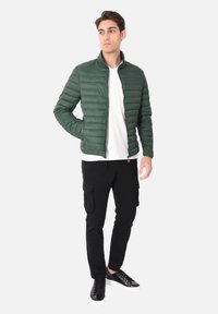 Giacca a vento verde con cuciture orizzontali, chiusura con zip e colletto alto, abbinata a pantaloni cargo neri e sneaker neri.