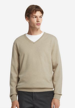Beige V-Ausschnitt-Pullover aus weichem Stoff. Mit gerippten Bündchen und Saum, über einem weißen Rundhals-Shirt getragen. Einfaches Design und neutrale Farbe.
