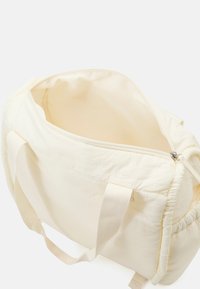 Sac de voyage en tissu crème avec une fermeture éclair sur le dessus, texture douce et anses plates tissées. L'intérieur est sobre, offrant un espace de rangement généreux.