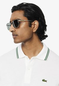 Polo shirt blanc avec un col à rayures vertes, comportant un petit logo brodé sur la poitrine. Modèle portant des lunettes de soleil sombres, tourné vers la gauche.