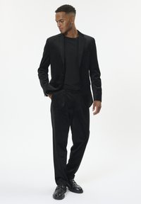 Homme portant un costume en velours noir, un t-shirt noir et des chaussures en cuir noires, se tenant avec une main dans la poche, regardant vers le bas sur le côté.