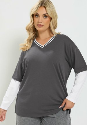 Donna bionda che indossa una maglietta a maniche corte grigio scuro con scollo a V sopra una maglia bianca a maniche lunghe e pantaloni grigi, in posa con la mano in tasca.