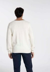 Pull en tricot blanc à manches longues, avec des poignets et un ourlet côtelés. Présente une encolure contrastante en noir et orange, texture lisse.