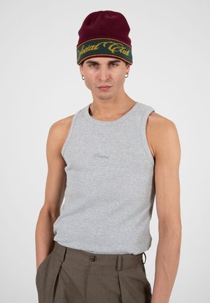 Mann iført en burgunderlue med gulgrønn tekst, grå ermeløs skjorte og brune plissert bukser mot en ensfarget lys bakgrunn.