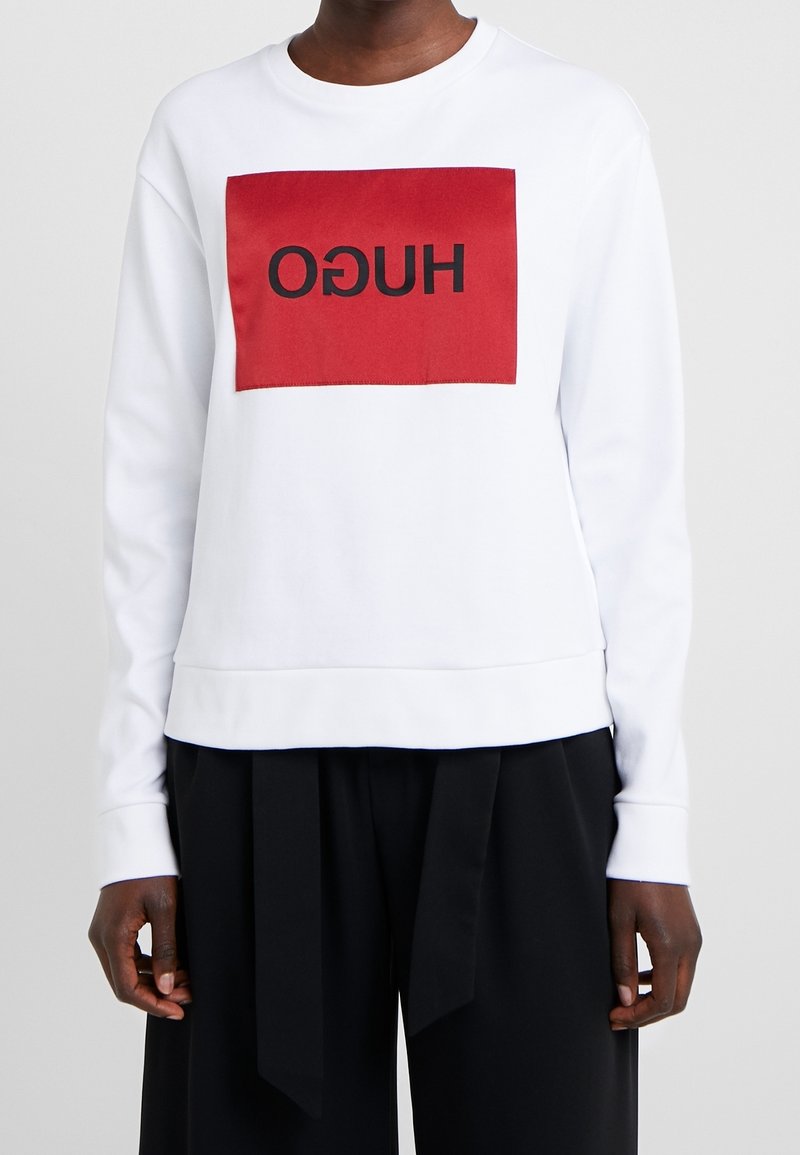 Personne portant une chemise blanche à manches longues avec un carré rouge et un texte noir inversé "HUGO" sur le devant, associée à un pantalon noir ample.