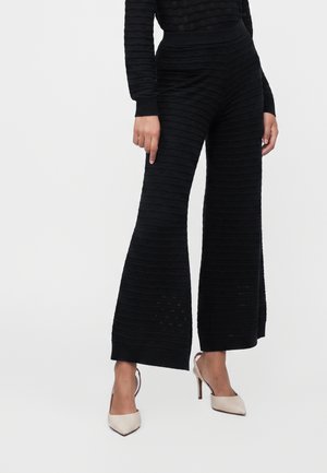 Femme portant un pantalon noir à jambes larges texturé et des chaussures à talons hauts beige à bout pointu, se tenant devant un fond uni.