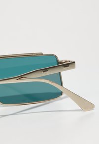 Occhiali da sole in metallo dorato con lenti color teal. Design della montatura quadrata con logo inciso sul lato. Stegole lunghe e sottili.