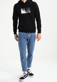 Persona che indossa un hoodie nero con scritte grafiche, jeans blu, calzini bianchi e sneaker bianco e nero, in piedi davanti a uno sfondo bianco semplice.