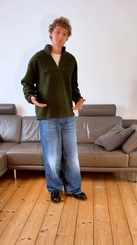 Pulover iz flisa olivno zelene barve z delnim zadrgo, kombiniran s svetlo modrimi širokimi kavbojkami in sijočimi črnimi čevlji, na minimalističnem ozadju.