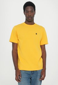 Tricou din bumbac galben, cu guler rotund, mâneci scurte și un mic logo negru pe piept. Purtați cu blugi din denim albastri.
