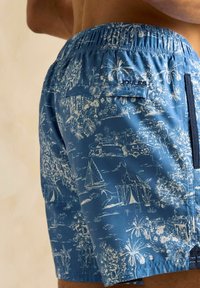 Șorturi de înot albastre cu modele tropicale albe, având un talie elastică, buzunare laterale și un buzunar posterior cu închidere tip clapetă.