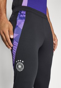 Pantaloni sportivi neri con un pannello laterale geometrico viola e un logo bianco che presenta un'aquila e cinque stelle. Texture liscia, design aderente.