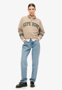 Sweatshirt beige avec l'inscription verte "SUPERDRY", col zippé et poignets rayés, associé à un jean bleu clair et des chaussures noires.