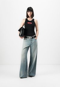 Frau mit schwarzem Bob-Haarschnitt, die ein schwarzes Tanktop und weite, ausgewaschene Jeans trägt, hält eine schwarze Clutch-Handtasche vor einem weißen Hintergrund.