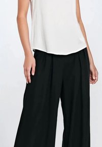 Biała bezrękawnikowa bluzka z zaokrąglonym dołem, połączona z czarnymi spodniami o szerokich nogawkach z wysokim stanem. Gładki materiał z detalami w formie fałd.