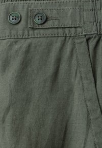 Gros plan sur un pantalon en tissu vert olive avec deux boutons vert foncé et une poche latérale avec des coutures visibles.