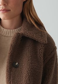 Veste d'extérieur brun, texturée, avec un grand col, fermeture à un bouton, et un tissu doux et pelucheux. Superposée sur un pull beige.