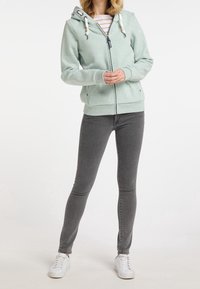 Světle zelená mikina na zip s kapucí na šňůrku, zdobená vzorovanými šňůrkami a bočními kapsami, ve spojení se šedými skinny džínami a bílými teniskami.