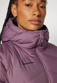 Lila puffjacka med hög krage, dragkedjestängning och svart "MAMMUT"-logotyp på bröstet. Har en slät, quiltad textur.