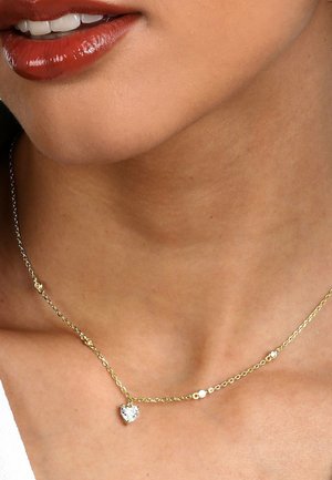 Nahaufnahme eines Frauenthalses mit glänzend roten Lippen und einer filigranen goldenen Kette mit einem kleinen herzförmigen Anhänger.