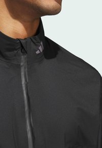 Svart lättviktsjacka med hög krage, försedd med dragkedja och en liten lila logotyp på kragen. Slät, matt tygstruktur.