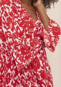 Robe à motif rouge et blanc avec un décolleté en V, des manches évasées et un tissu texturé. Présente un design fluide et des plis visibles.