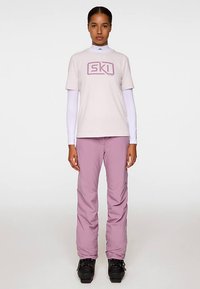 Ljusrosa skid-t-shirt med "SKI"-logotyp, kombinerad med lila skidbyxor. Outfitten har en figurnära design och långa vita underarmar.