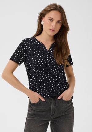 Femme aux longs cheveux bruns porte une chemise noire à manches courtes à pois et un jean foncé, debout les mains dans les poches devant un fond uni.