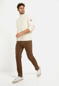 Pull à col roulé crème côtelé avec un accent en cuir sur l'épaule, associé à un pantalon en velours côtelé marron et des baskets blanches.