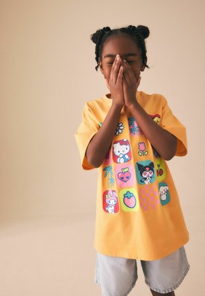 Kind mit geflochtenem Haar, das ein gelbes T-Shirt mit Cartoon-Motiv und helle Shorts trägt, bedeckt das Gesicht mit den Händen und steht vor einem beigen Hintergrund.