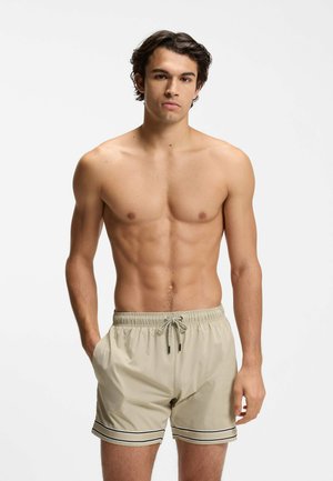 Giovane con capelli scuri che indossa pantaloncini da bagno beige con bordi neri, in piedi con una mano in tasca contro uno sfondo semplice.