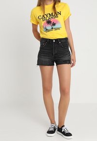 Yellow graphic t-shirt with "CAYMAN ISLANDS" text and palm tree design, paired with high-waisted black denim shorts and black sneakers.