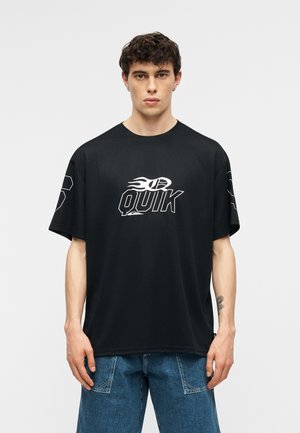 Jeune homme aux cheveux courts et bouclés portant un T-shirt noir oversized avec le logo "QUIK" et un pantalon en denim bleu, debout devant un fond clair uni.