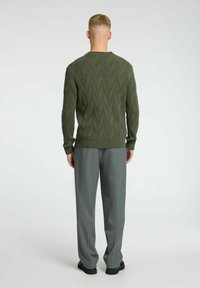 Olive spalvos megztinis su tekstūruotu lapų raštu, derinamas su šviesiai pilkais pritaikytais kelnėmis ir juodais batais.