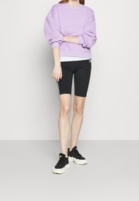 Kvinna som bär en lavendelfärgad sweatshirt, svarta Adidas cykelshorts med vita ränder, och svart-vita Adidas sneakersstående mot en vit vägg.