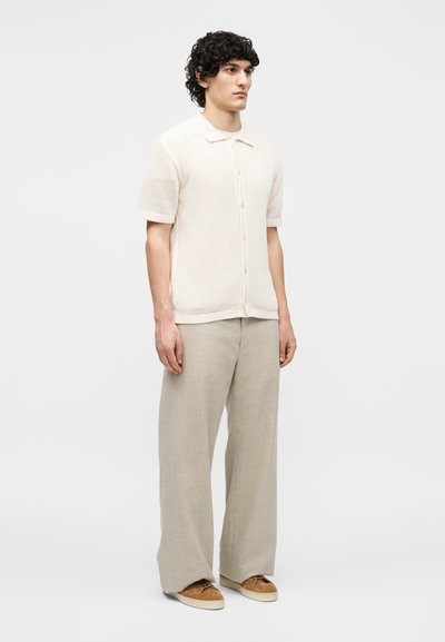 Άνδρας που στέκεται φορώντας μια κρεμ πλεκτή κοντομάνικη μπλούζα, μπεζ παντελόνι με φαρδιά μπατζάκια και καφέ casual παπούτσια σε λευκό φόντο.