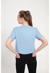Světle modré tričko s krátkým rukávem a kulatým výstřihem, hladká textura, volný střih, nošeno s černými džíny, viděno zezadu.