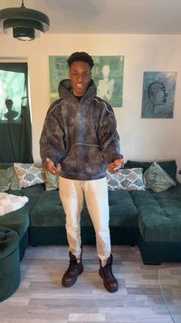 Bărbat tânăr purtând un hanorac închis la culoare și pantaloni deschiși, stând într-un living cu o canapea verde secțională și artă abstractă pe perete în spatele lui.