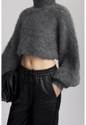 Personne portant un pull court à col roulé gris pelucheux et un pantalon en cuir noir, tenant une pochette noire, main dans la poche.