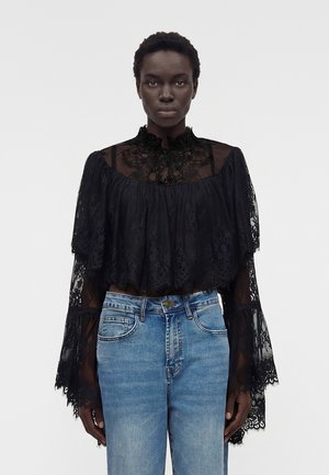 Γυναίκα που φοράει μια διαφανή μαύρη δαντελένια μπλούζα με βολάν στον ψηλό λαιμό και φαρδιά μανίκια, συνδυασμένη με ανοιχτό μπλε τζιν με ψηλή μέση.