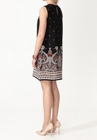 Kvinna i en ärmlös svart klänning med intrikata vita och röda paisleymönster i kanten, nakna högklackade skor och pärlarmband, stående på sidan.