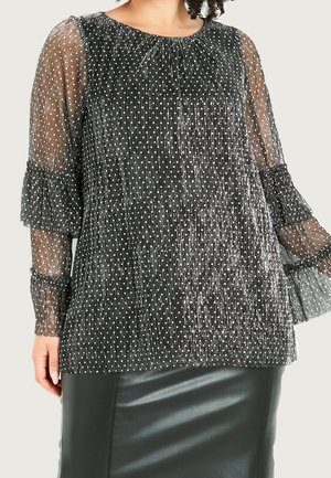 Blusa negra transparente con lunares blancos, tela plisada, mangas largas con volantes, combinada con una falda de cuero negra.