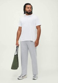 White cotton t-shirt, gray textured sweatpants, and white sneakers. Holding a green quilted tote bag. Standing on a neutral background.